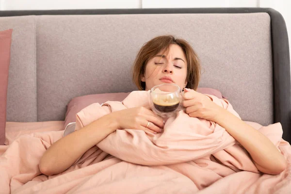Young sleepy woman lying on the bed and drinking hot coffee for waking up and gaining energy