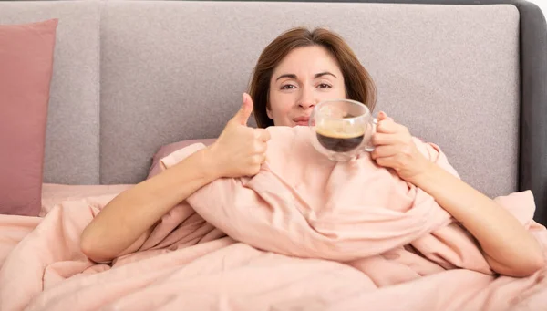 年轻的睡意朦胧的女人躺在床上 喝着热咖啡醒来 精力充沛 — 图库照片
