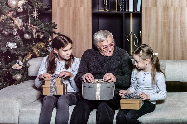 Little granddaughters and their grandpa sitting on the couch, trying to open gift boxes and celebrating Christmas together