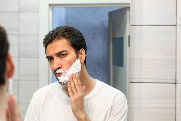 Handsome man looking at the mirror and shaving in the bathroom