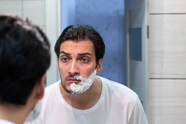 Handsome man looking at the mirror and shaving in the bathroom