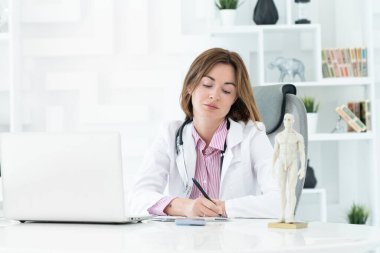 Modern kliniğin ofisinde dizüstü bilgisayarda oturan ve çalışan beyaz elbiseli, gülümseyen genç ve güzel bir kadın doktorun portresi. Çevrimiçi danışma ve uzak tedavi kavramı