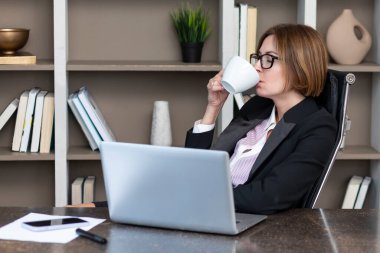 Kendine güvenen ve çekici bir iş kadını. Masada çalışıyor ve modern ofiste kahve fincanı tutuyor.