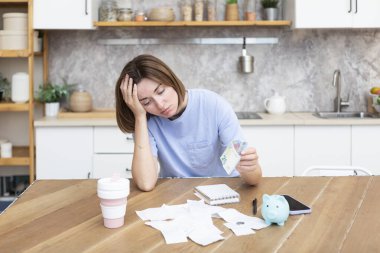 Unhappy and depressed woman sitting in the kitchen at the table, holding piggy bank and looking at a lot of bills and checks. Taxes and debts, family budget and spending concept