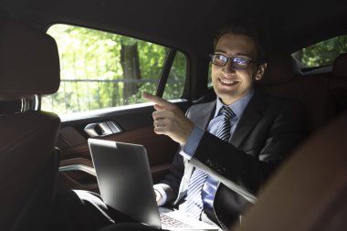 Başarılı yakışıklı işadamı lüks arabasında yolcu koltuğunda oturuyor dizüstü bilgisayarla çalışıyor ve bir iş toplantısına gidiyor.