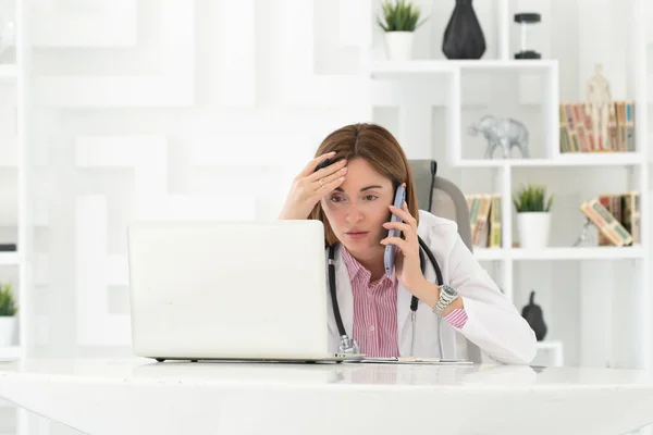 Close up portrait of tired female doctor sitting at the desktop and talking to phone in the office of modern clinic
