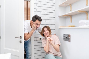 Genç heyecanlı kadın tuvalette oturuyor ve kocasına hamilelik testinin sonucunu gösteriyor.