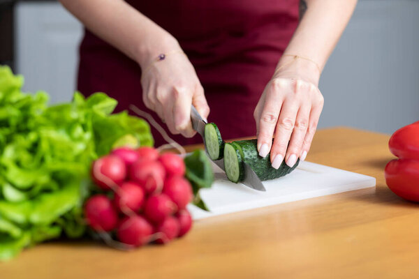 Молодая привлекательная женщина в красном фартуке нарезает огурец и готовит вегетарианскую еду