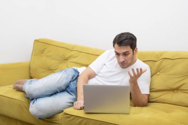 Kanepede oturan yakışıklı adam evde dizüstü bilgisayarla çalışıyor. Çevrimiçi eğitim ve uzak çalışma kavramı