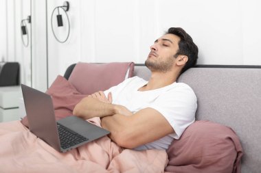 Evde dizüstü bilgisayarla çalışırken yatakta uyuyan tembel genç yakışıklı adam.