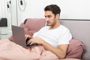 Yakışıklı gülümseyen adam yatakta yatar ve laptopla yatak odasında çalışır.