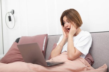 Yatakta dizüstü bilgisayarla çalışan elleri başında şok olmuş bir kadın. Şaşırmış ve üzgün kız yatak odasındaki bilgisayarına bakıyor.