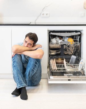 Mutfaktaki bulaşık makinesine kirli tabaklar koymaktan yorgun ve sıkılmış bir adam. Ev yapımı ve yorucu temizlik günü konsepti