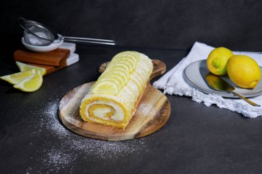 İçinde limon kremalı vanilyalı kek rulosu, arka planda şeker tozu ve limonlu gri mermer masa.