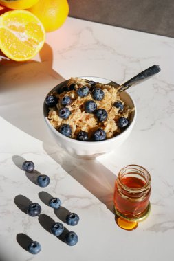 healthy oatmeal with blueberries on top in white porcelain bowl, honey jar and oranges in background . White marble table and grey wall 