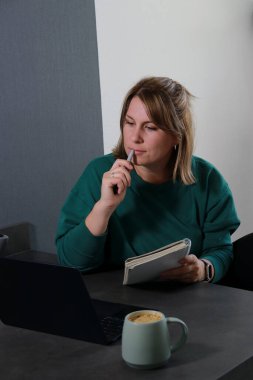 Dizüstü bilgisayarda çalışan genç bir kadın ve derin düşüncelere dalmış. Not defteri ve kahve fincanı masanın üstünde..