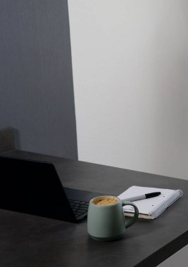 Çalışma alanı yeşil kahve fincanı, defter ve gri mermer masada siyah dizüstü bilgisayar.