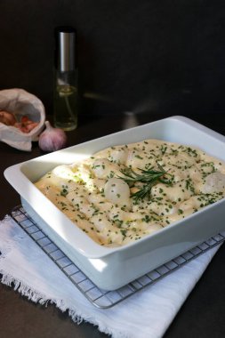 Çiğ Focaccia hamuru, sarımsaklı biberiyeli, koyu gri masalı, beyaz fırın kabında.