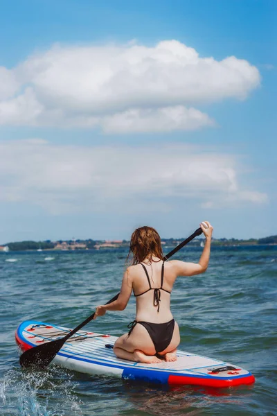 ブロンドの髪の女性の背中のビューは 海の中でパドルボードの上に立つ乗馬 夏の楽しいスポーツ活動 — ストック写真