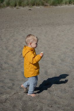 İki yaşında, kumsalda, sarı ceketli, güneşli bulutlu bir çocuk..