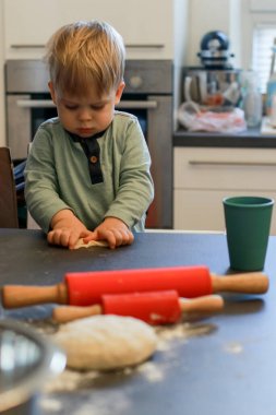 Küçük bir çocuğun yoğurt hamurunun üst görüntüsü, masada kırmızı oklavalar, arka planda dağınık bir mutfak.