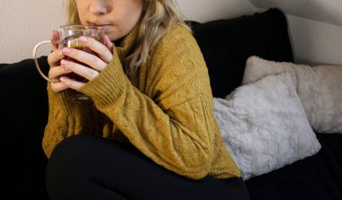 Limonlu ve ballı sıcak çay içen, koltukta oturan ve sarı kazak giyen bir kadın manzarası.