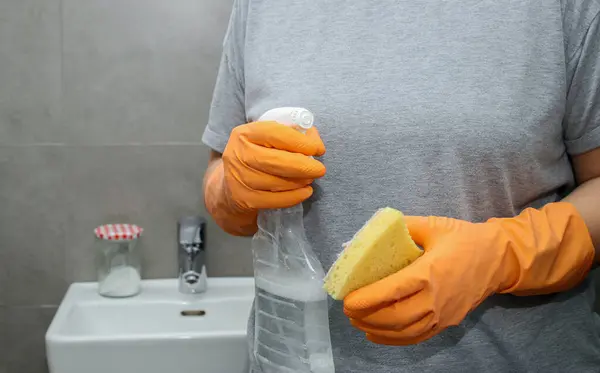 Close up of womans hand in orange gloves holding a yellow sponge and a cleaning spray bottle.