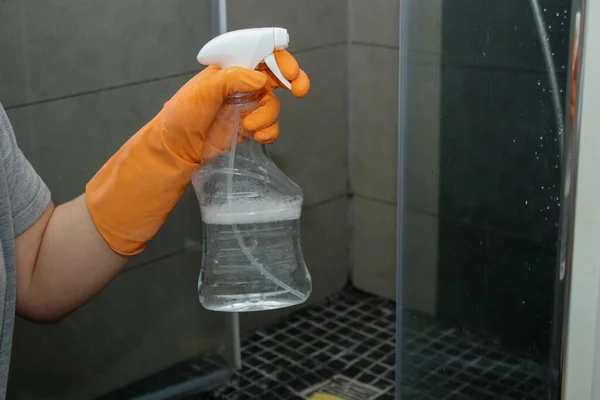 Close up of womans hand in orange gloves cleaning dirty glass shower doors with lime remover spray