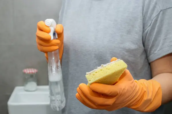 Close up of womans hand in orange gloves holding a yellow sponge and a cleaning spray bottle.