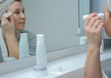 Bornoz ve havlu giymiş genç bir kadın banyodaki aynaya bakıyor, göz bakımı uyguluyor.. 