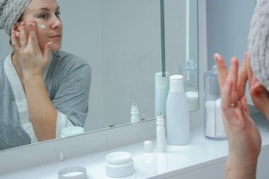 Bornoz ve havlu giymiş genç bir kadın banyodaki aynaya bakıyor, cilt kremi sürüyor.. 