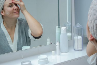 Bornoz ve havlu giymiş genç bir kadın banyodaki aynaya bakıyor, cilt kremi sürüyor.. 