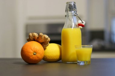 Ev yapımı portakal, limon ve zencefilli içeceği mutfak masasında küçük bardakta, bulanık arka planda bir cam şişeye koy. 