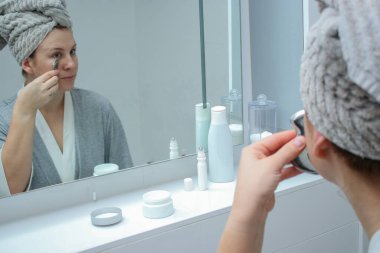 Bornoz ve havlu giymiş genç bir kadın banyodaki aynaya bakıyor, metal bir guashayla yüz masajı yapıyor.. 