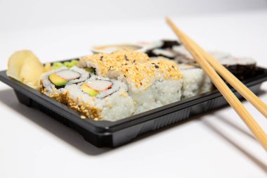 Suşi rulolarını kapayın ve siyah bir yemek kutusuna koyun. Beyaz arka planda tahta çubuklar olsun. 