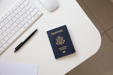 Beyaz masadaki siyah tavalı, klavyesi ve faresi olan bir Amerikan pasaportunun üst görüntüsü.