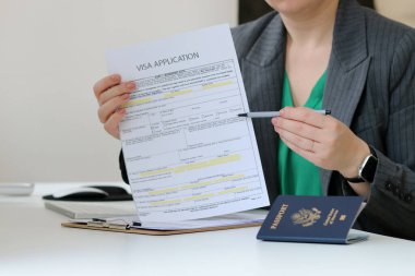 Kafkasyalı bir kadının elinde vize başvuru kâğıdı belgesi ve ofis masasında Amerikan pasaportu olan vize başvurusu konseptini yakın plan.