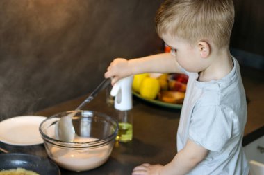 Mutfakta krep pişiren beyaz bir çocuk. Masada hamurlu cam kase ve diğer malzemeler var. 