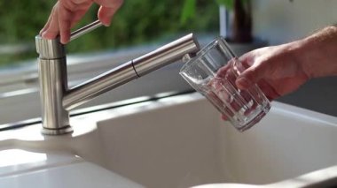 Erkek ellerinin mutfaktaki bardağa musluk suyu dökmesini kapat. Beyaz lavabo ve bulanık arkaplan 