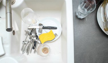 Beyaz seramik mutfak lavabosu kirli bulaşıklarla dolu, koyu gri masa.