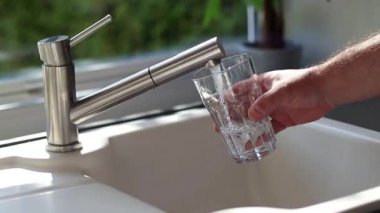 Erkek ellerinin mutfaktaki bardağa musluk suyu dökmesini kapat. Beyaz lavabo ve bulanık arkaplan 