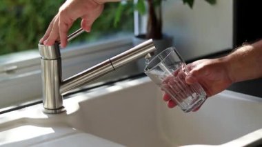Erkek ellerinin mutfaktaki bardağa musluk suyu dökmesini kapat. Beyaz lavabo ve bulanık arkaplan 