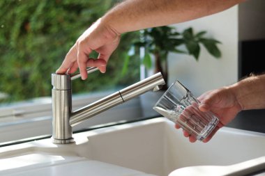 Erkek ellerinin mutfaktaki bardağa musluk suyu dökmesini kapat. Beyaz lavabo ve bulanık arkaplan 