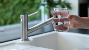 Kadın ellerinin musluk suyuyla bardağı tutuşu, arka planda bulanık bir lavabo.