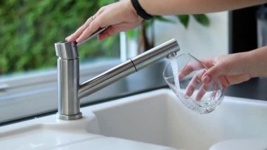 Kadın ellerinin mutfaktaki bir bardağa musluk suyu dökmesini kapatın. Beyaz lavabo ve bulanık yeşil arka plan. 