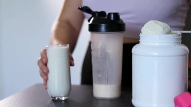 Bardakta protein içeceği içen bir kadının yakın çekimi. Kilo vermenin yerine diyet yemeği. Masada beyaz beyaz protein tozu ve pembe dambıllar var. 