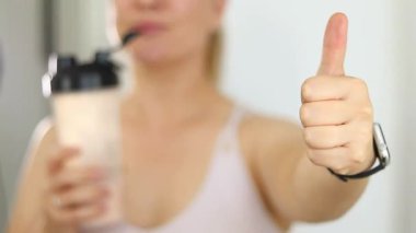 Protein içeceği içen ve başparmağını kaldıran bir kadının yakın çekimi. Kilo vermenin yerine diyet yemeği. Masada beyaz beyaz protein tozu ve pembe dambıllar var. 