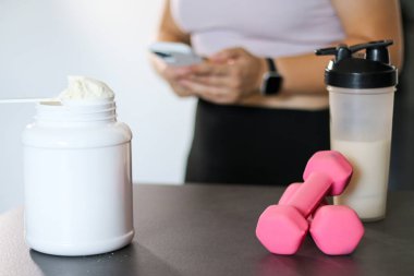 Bir protein içeceği ve sehpanın üzerindeki dambılları kapatın, arka planda akıllı telefonuyla kalori alan bir kadın. Kilo kaybı için diyet yemeği.