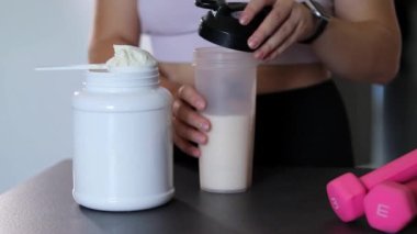 Mutfakta protein içeceği hazırlayan kadına yakın çekim. Kilo vermenin yerine diyet yemeği. Masada beyaz beyaz protein tozu ve pembe dambıllar var. 