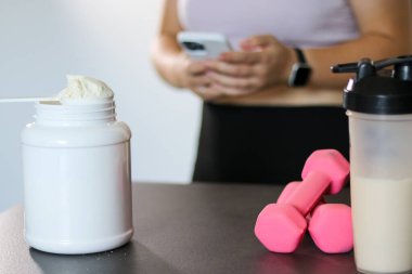 Bir protein içeceği ve sehpanın üzerindeki dambılları kapatın, arka planda akıllı telefonuyla kalori alan bir kadın. Kilo kaybı için diyet yemeği.